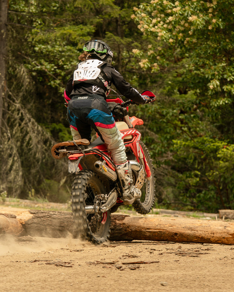 Person riding a dirt bike over a log in a forest setting