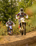 Two dirt bike riders on a trail with trees and flowers in the background