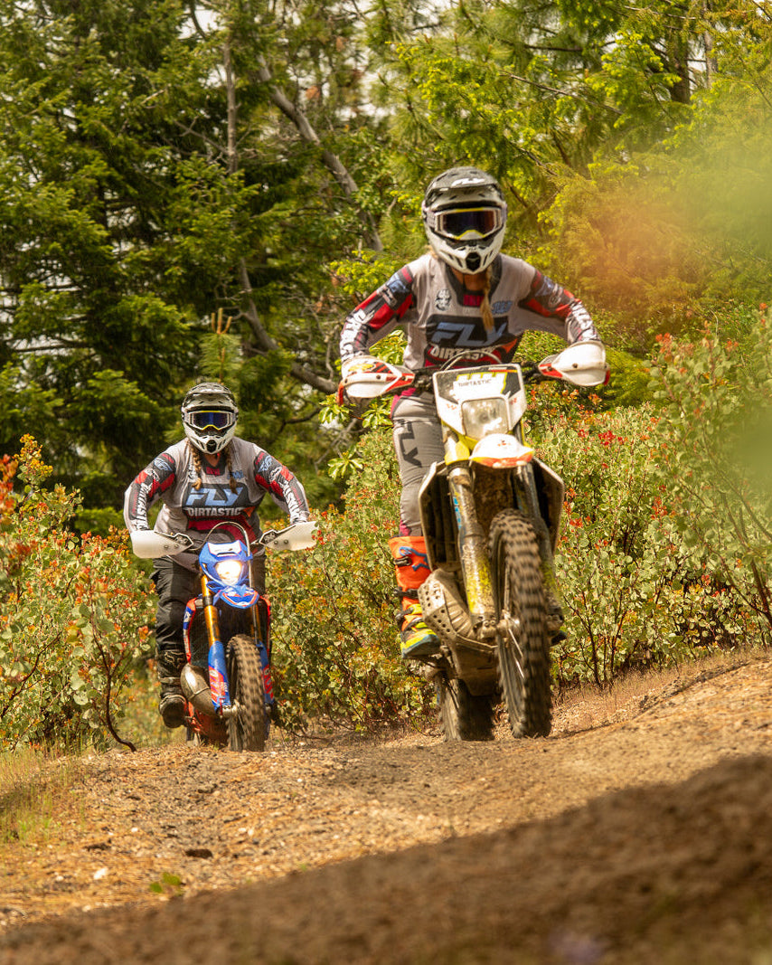 Two dirt bike riders on a trail with trees and flowers in the background