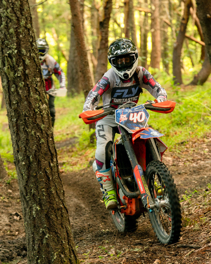 Two dirt bikers navigating a forest trail with trees and greenery in the background.